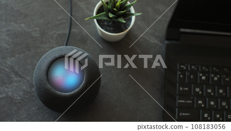Using an HomePod Mini speaker - the smart speaker is reacting to voice listening to human commands and signalling its attention by the changing lights. Concept of a smart home. Using an HomePod Mini speaker - the smart speaker is reacting to voice listening to human commands and signalling its attention by the changing lights. Concept of a smart home. 108183056