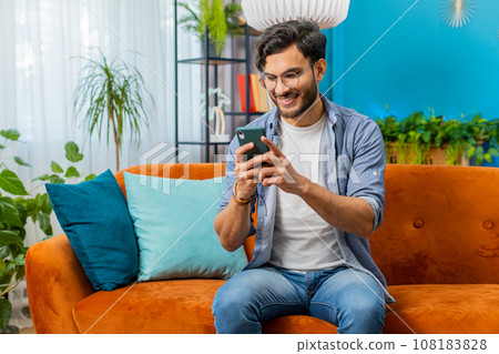 Indian Arabian young man sitting on sofa use smartphone share messages on social media application 108183828