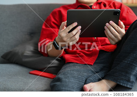 Relaxed young man sitting on couch and browsing internet or watching social media videos on digital tablet Relaxed young man sitting on couch and browsing internet or watching social media videos on digital tablet 108184365