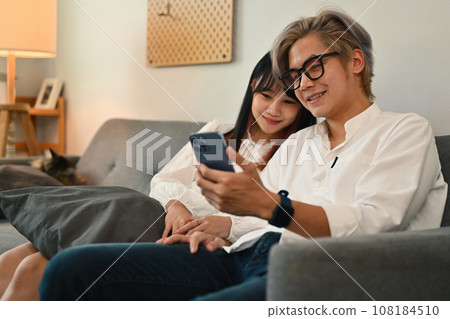 Attractive young couple spending weekend time on couch in living room using smartphone together 108184510