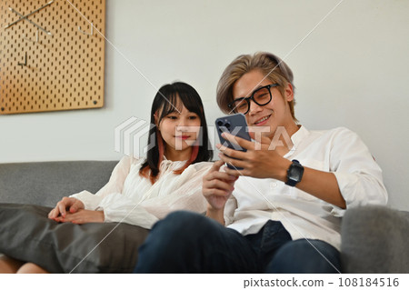 Happy young couple sitting on sofa and watching social media videos on smartphone Happy young couple sitting on sofa and watching social media videos on smartphone 108184516