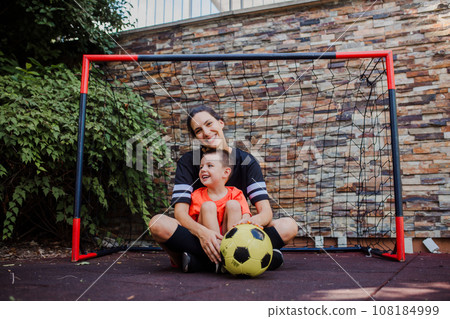 Mom playing football with her son, dressed in football jerseys. The family as one soccer team. Family sports activities outside in the backyard or on the street. 108184999
