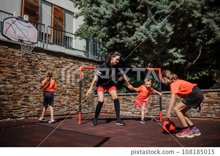 Mom playing football with her children, dressed in football jerseys. The family as one soccer team. Family sports activities outside in the backyard or on the street. 108185015