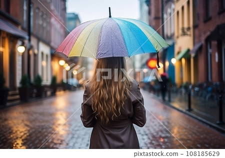 Girl walks in rain in gray city, carrying rainbow umbrella, view from back Girl walks in rain in gray city, carrying rainbow umbrella, view from back 108185629