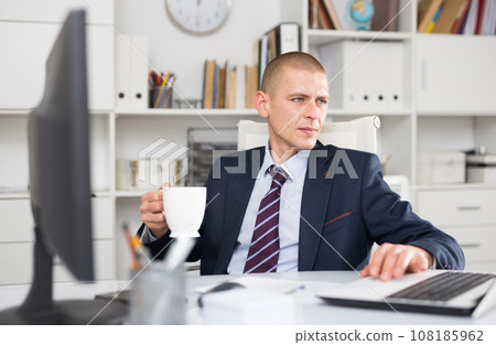 Male head of the company, working at a laptop, holding a cup of coffee in his hand 108185962