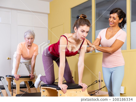 Young woman doing pilates on combo chair with Hispanic female trainer Young woman doing pilates on combo chair with Hispanic female trainer 108186334