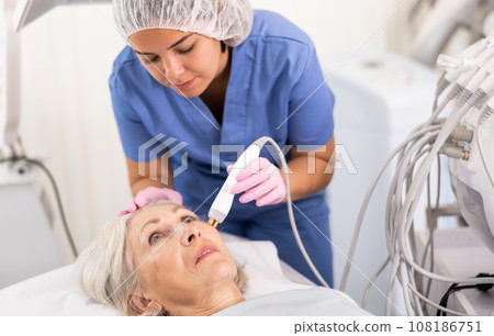 Elderly woman at appointment with beautician doing ultrasonic face cleaning with help of high-frequency ultrasonic waves in cosmetology clinic 108186751