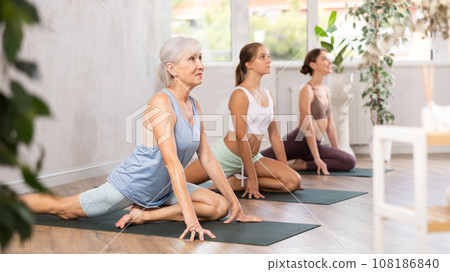 Three generations of women perform sitting pigeon pose Eka Pada Rajakapotasana in gym 108186840