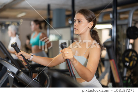 Slender athletic girl during cardio exercises on elliptical simulator in fitness center. Slender athletic girl during cardio exercises on elliptical simulator in fitness center. 108187275