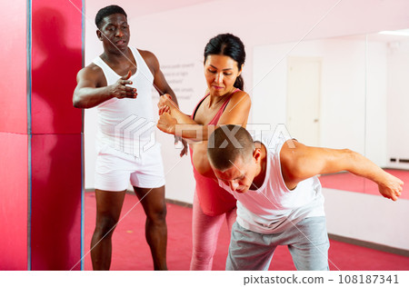 Asian woman doing strength painful hold while training self-defense in pairs with the help of trainer in gym 108187341