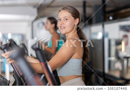 Slender athletic girl during cardio exercises on elliptical simulator in fitness center. Slender athletic girl during cardio exercises on elliptical simulator in fitness center. 108187448