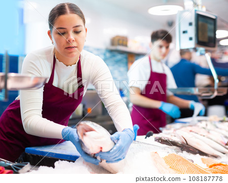 Complaisant female seller in apron standing near counter offering fresh fish atlantic bonito to buyer 108187778