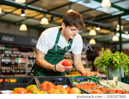 Focused young salesman laying out ripe tomatoes on display supermarket Focused young salesman laying out ripe tomatoes on display supermarket 108187860