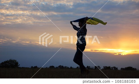 Happy lady walking on barley meadow with a raised over head flag of Ukraine. Ukrainian woman with a lifted blue-yellow banner on a beautiful sunset at background. End of war concept. 108188103