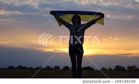 Happy lady walking on barley meadow with a raised over head flag of Ukraine. Ukrainian woman with a lifted blue-yellow banner on a beautiful sunset at background. End of war concept. 108188106