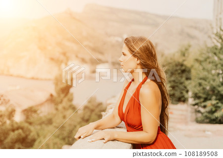Woman red dress. Summer lifestyle of a happy woman posing near a fence with balusters over the sea. Woman red dress. Summer lifestyle of a happy woman posing near a fence with balusters over the sea. 108189508