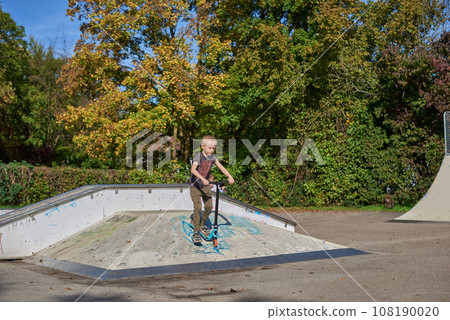 Boy on scooter makes a trick and enjoying his riding in the skate park at cloudy spring day. Young man doing trick on the kick scooter in the park. Full body little boy on the kick scooter on sunny 108190020