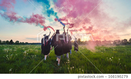 Graduates in costume walk across a field with coloured red and blue smoke at sunset. 108190279