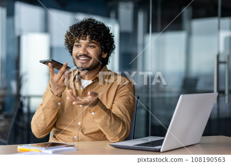 Cheerful and smiling successful businessman using app on phone to record audio message, Indian man inside office with laptop working. A financier talks on the phone, uses an assistant. Cheerful and smiling successful businessman using app on phone to record audio message, Indian man inside office with laptop working. A financier talks on the phone, uses an assistant. 108190563