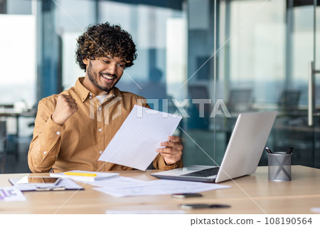 Portrait of young successful hispanic businessman financier, man smiling contentedly, holding hand up celebrating victory, and successful achievement results, investor paperwork. Portrait of young successful hispanic businessman financier, man smiling contentedly, holding hand up celebrating victory, and successful achievement results, investor paperwork. 108190564
