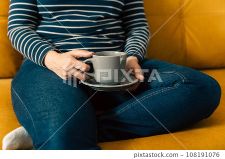woman in a striped sweater sits on the sofa and holds a cup of coffee in hands woman in a striped sweater sits on the sofa and holds a cup of coffee in hands 108191076
