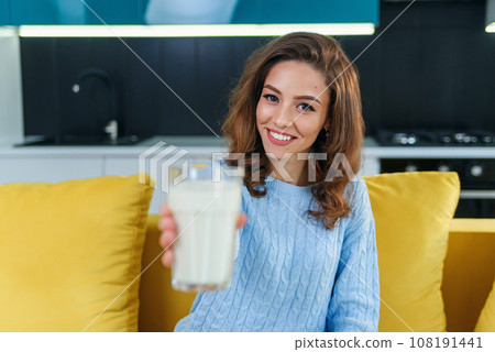 Smiling woman gives to the camera a glass of fresh milk in the stylish cozy kitchen at the morning. Pretty healthy girl holds a glass of fresh milk on foreground. 108191441