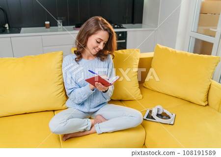 Likable young woman writing her personal notes into her diary while sitting in the modern stylish kitchen. Likable young woman writing her personal notes into her diary while sitting in the modern stylish kitchen. 108191589