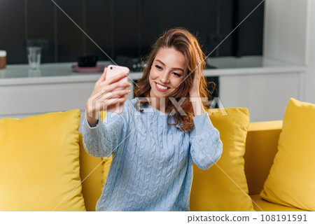 Attractive cheerful girl makes selfie photo in modern stylish kitchen. Young woman having fun on the yellow couch at cozy home. Attractive cheerful girl makes selfie photo in modern stylish kitchen. Young woman having fun on the yellow couch at cozy home. 108191591