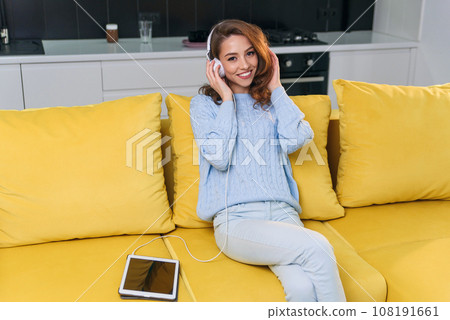 Adorable modern woman with wavy brown hair dressed in stylish casual clothes resting on the comfortable sofa in the kitchen and listens to music in headphones. Cozy home concept. 108191661
