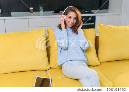 Adorable modern woman with wavy brown hair dressed in stylish casual clothes resting on the comfortable sofa in the kitchen and listens to music in headphones. Cozy home concept. 108191662