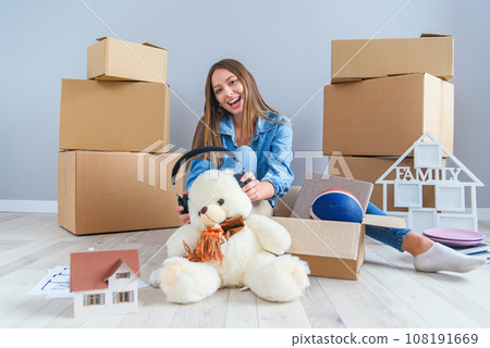 Beautiful smiling girl puts on the headphones on the teddy bear toy and sitting on the floor in the empty new apartment. 108191669