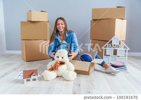 Beautiful smiling girl puts on the headphones on the teddy bear toy and sitting on the floor in the empty new apartment. 108191670