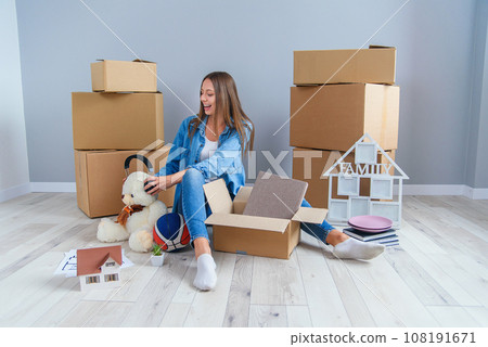 Beautiful smiling girl puts on the headphones on the teddy bear toy and sitting on the floor in the empty new apartment. 108191671