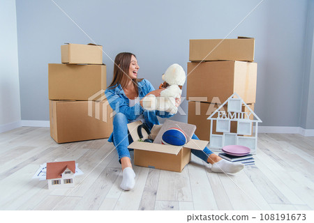 Beautiful smiling girl puts on the headphones on the teddy bear toy and sitting on the floor in the empty new apartment. Beautiful smiling girl puts on the headphones on the teddy bear toy and sitting on the floor in the empty new apartment. 108191673