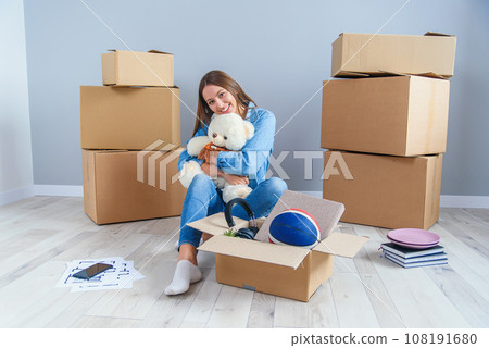 Beautiful smiling girl puts on the headphones on the teddy bear toy and sitting on the floor in the empty new apartment. 108191680