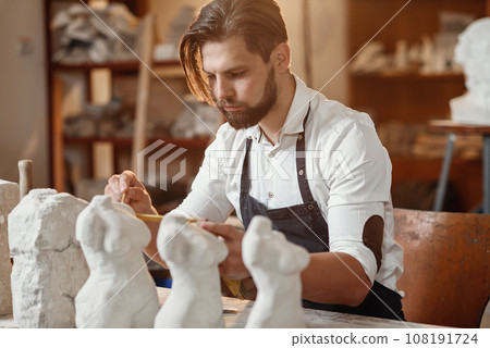 Concentrated 30-aged creative bearded stonemason knocks off from limestone needed shape of future sculpture using wooden hammer and iron chisel. Concentrated 30-aged creative bearded stonemason knocks off from limestone needed shape of future sculpture using wooden hammer and iron chisel. 108191724