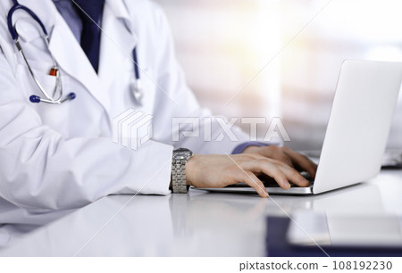 Unknown male doctor sitting and working with laptop in a darkened clinic, glare of light on the background, close-upof hands 108192230
