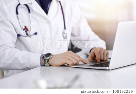 Unknown male doctor sitting and working with laptop in a darkened clinic, glare of light on the background, close-upof hands 108192239