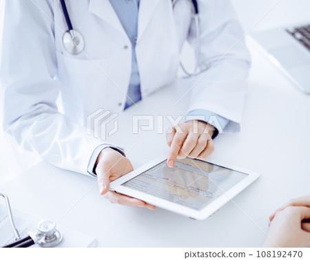 Doctor and patient sitting opposite each other at the desk in clinic. The focus is on female physician's hands pointing into tablet computer touchpad, close up. Medicine concept Doctor and patient sitting opposite each other at the desk in clinic. The focus is on female physician's hands pointing into tablet computer touchpad, close up. Medicine concept 108192470