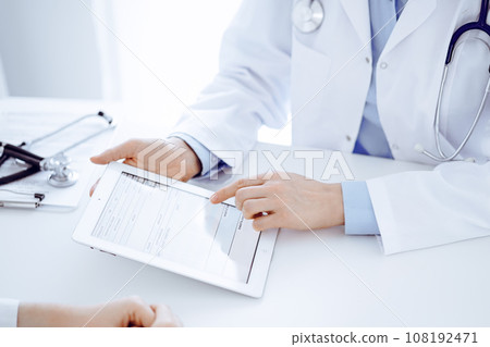 Doctor and patient sitting opposite each other at the desk in clinic. The focus is on female physician's hands pointing into tablet computer touchpad, close up. Medicine concept Doctor and patient sitting opposite each other at the desk in clinic. The focus is on female physician's hands pointing into tablet computer touchpad, close up. Medicine concept 108192471