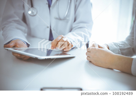 Doctor and patient sitting near each other at the desk in clinic. The focus is on female physician's hands pointing into tablet computer touchpad, close up. Medicine concept Doctor and patient sitting near each other at the desk in clinic. The focus is on female physician's hands pointing into tablet computer touchpad, close up. Medicine concept 108192489