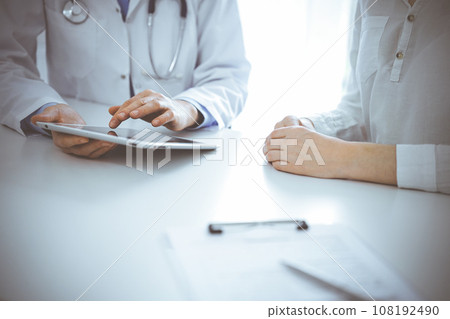 Doctor and patient sitting near each other at the desk in clinic. The focus is on female physician's hands pointing into tablet computer touchpad, close up. Medicine concept Doctor and patient sitting near each other at the desk in clinic. The focus is on female physician's hands pointing into tablet computer touchpad, close up. Medicine concept 108192490