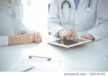 Doctor and patient sitting near each other at the desk in clinic. The focus is on female physician's hands pointing into tablet computer touchpad, close up. Medicine concept Doctor and patient sitting near each other at the desk in clinic. The focus is on female physician's hands pointing into tablet computer touchpad, close up. Medicine concept 108192491