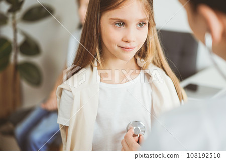 Doctor and kid patient are in the clinic. Physician in white coat examining a serious young girl with a stethoscope, close up. Medicine, therapy concept Doctor and kid patient are in the clinic. Physician in white coat examining a serious young girl with a stethoscope, close up. Medicine, therapy concept 108192510