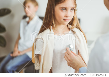 Doctor and kid patient are in the clinic. Physician in white coat examining a serious young girl with a stethoscope, close up. Medicine, therapy concept Doctor and kid patient are in the clinic. Physician in white coat examining a serious young girl with a stethoscope, close up. Medicine, therapy concept 108192511
