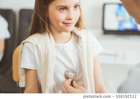 Doctor and kid patient are in the clinic. Physician in white coat examining a smiling young girl with a stethoscope, close up. Medicine, therapy concept Doctor and kid patient are in the clinic. Physician in white coat examining a smiling young girl with a stethoscope, close up. Medicine, therapy concept 108192512