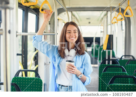 Stylish woman in blue shirt enjoying trip in the modern tram or bus, stands with cup of coffee in the public transport. Stylish woman in blue shirt enjoying trip in the modern tram or bus, stands with cup of coffee in the public transport. 108193487