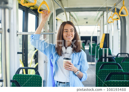 Stylish woman in blue shirt enjoying trip in the modern tram or bus, stands with cup of coffee in the public transport. Stylish woman in blue shirt enjoying trip in the modern tram or bus, stands with cup of coffee in the public transport. 108193488