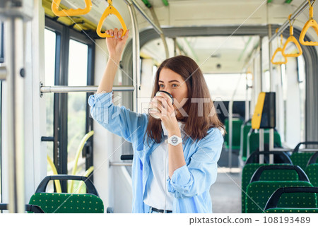 Beautiful woman in blue shit drinks coffee while riding in the public transport. Beautiful woman in blue shit drinks coffee while riding in the public transport. 108193489