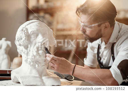 Professional bearded carver polishing with micro drill machine details of the man's sculpture head in his workshop. Professional bearded carver polishing with micro drill machine details of the man's sculpture head in his workshop. 108193512